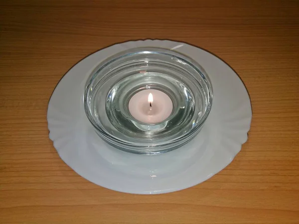 A tealight candle placed in a glass bowl and placed on a white porcelain plate, all on a wooden board, image