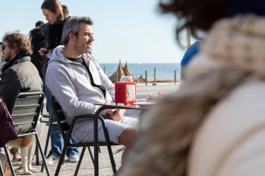 Barselona, İspanya - Jan 08, 2022: Bir adam terasta oturuyor, güneşlenirken biriyle sohbet ediyor ve Barceloneta plajının önünde garsonların servis yapmasını bekliyor