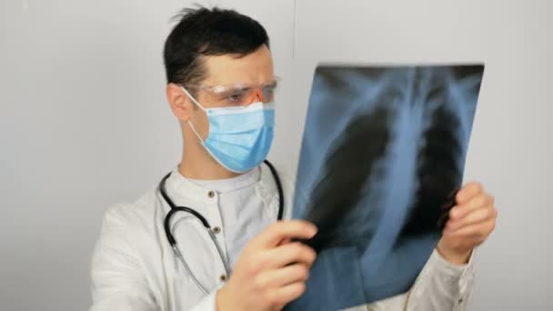 Młody chirurg noszący ochronną maskę medyczną bada prześwietlenie płuc pacjenta i rozważa diagnozę. Młody przystojny lekarz w masce medycznej trzyma zdjęcie rentgenowskie płuc w swoim — Wideo stockowe