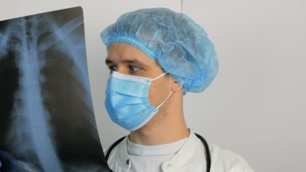 Молодой хирург в защитной медицинской маске исследует рентген легких пациента и размышляет над диагнозом. — стоковое видео