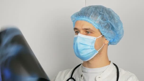 Egy fiatal sebész védőmaszkot visel, megvizsgálja a beteg tüdejét, és elgondolkodik a diagnózison. Egy fiatal, jóképű orvos orvosi maszkban röntgenképet tart a tüdejéről. — Stock videók