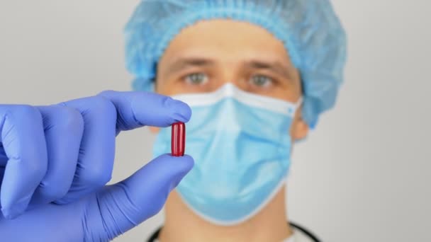 Przystojny młody lekarz bada nowy lek. Młody przystojny lekarz w masce medycznej trzyma w rękach czerwoną pigułkę przedstawiającą nowy lek.. — Wideo stockowe