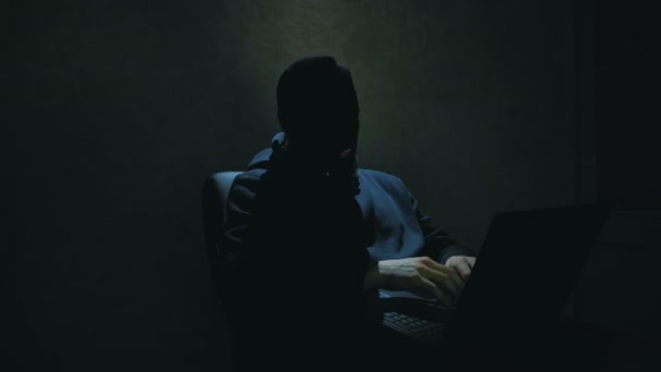 Een anonieme hacker in een zwarte hoodie krakt wachtwoorden en downloadt belangrijke databases van internetgebruikers. Een internet hacker in een zwarte bivakmuts zit in een stoel en steelt data van een laptop — Stockvideo