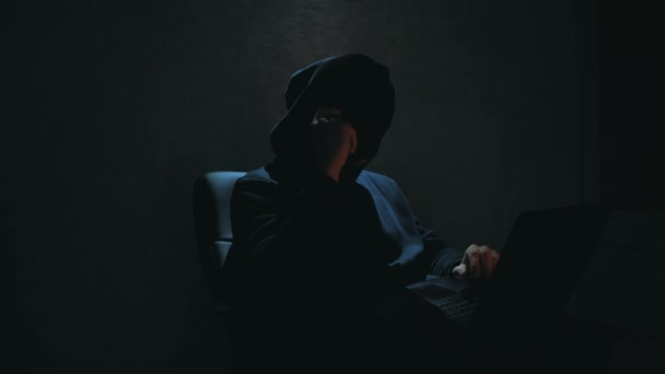 Um hacker anónimo com um capuz preto liga para a vítima ao telefone e ameaça decifrar as senhas da base de dados. Um hacker de Internet em uma balaclava preta senta-se em uma cadeira e rouba dados de um laptop — Vídeo de Stock