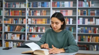 Genç çekici kız öğrenci bir kitap okuyor ve bir not defterine not alıyor, bir halk kütüphanesinde kitaplıkların arka planında otururken sınav için hazırlanıyor. Eğitim konsepti.