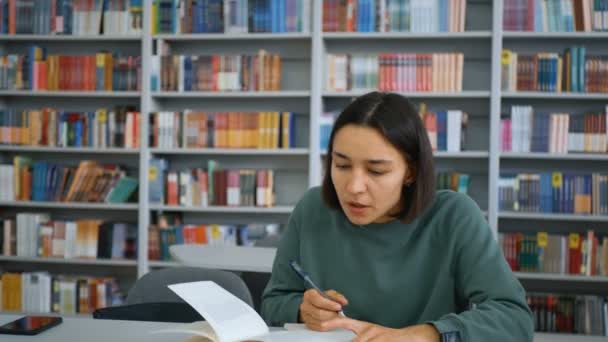 Die junge attraktive Studentin liest ein Buch, macht sich Notizen in einem Notizbuch und bereitet sich an einem Tisch in einer öffentlichen Bibliothek vor dem Hintergrund von Bücherregalen auf eine Prüfung vor.. — Stockvideo