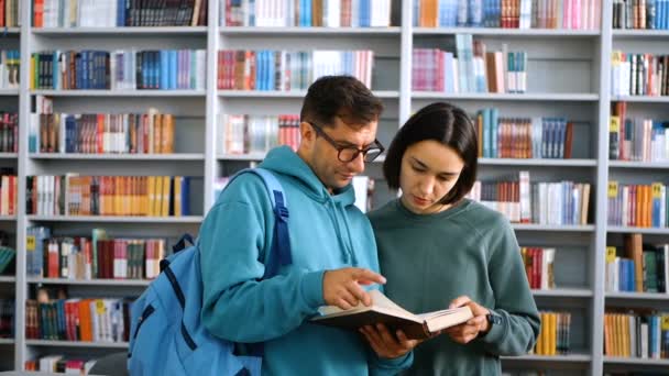 Una joven estudiante milenaria con gafas y una joven atractiva estudiante están leyendo un libro, preparándose para un examen mientras están de pie en una biblioteca pública en el contexto de las estanterías. concepto. — Vídeos de Stock