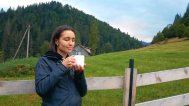 Genç çekici kadın kahve içiyor ve dağların ve tarlanın güzel manzarasının tadını çıkarıyor. Genç çekici kadın çiftçi, çiftliğinin ahşap çitlerinin yanında dururken kahve içer..