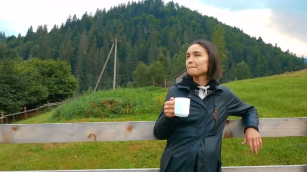 若い魅力的な女性はコーヒーを飲んでいて、山と畑の美しい景色を楽しんでいます。若い魅力的な女性の農家は彼女の牧場の木製の柵のそばに立っている間コーヒーを飲んでいます — ストック動画