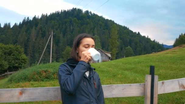 매력적 인 젊은 여자가 코 오피 를 마시면서 산 과들의 아름다운 경치를 즐기고 있다. 사랑 스러운 한 젊은 여자 농부 가 목장 의나무 울타리 옆에서 서 향기 로운 커피를 즐긴다. — 비디오