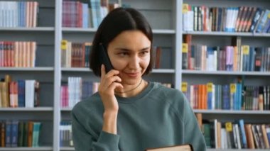 Genç bir milenyum kadını kütüphanede kitaplıkların önünde dururken cep telefonuyla konuşuyor..