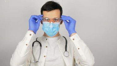 Genç doktor hastayı nazikçe selamlayarak elini sallıyor. Genç yakışıklı doktor hastaları muayene etmeye hazırlanırken güvenlik gözlüğü ve tıbbi maske takıyor..