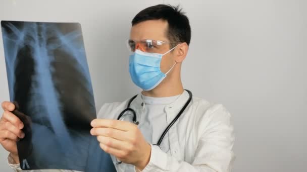 Młody chirurg w ochronnej masce medycznej bada prześwietlenie płuc pacjenta i rozważa diagnozę. — Wideo stockowe