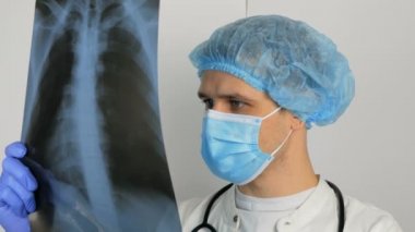 Koruyucu tıbbi maske takan genç bir cerrah hastaların akciğerlerinin röntgenini inceler ve teşhisi düşünür. Tıbbi maskeli genç ve yakışıklı bir doktor ciğerlerinde röntgen filmi tutuyor.