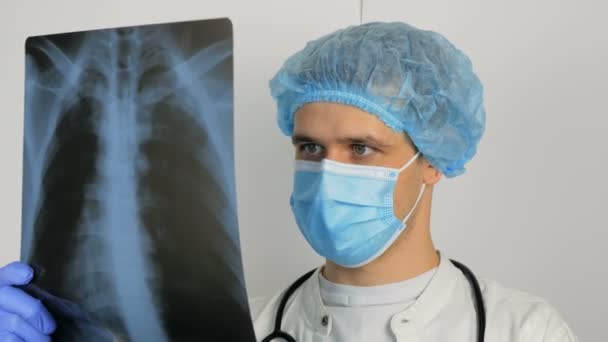 Un giovane chirurgo che indossa una maschera protettiva medica esamina una radiografia di un polmoni pazienti e medita la diagnosi. Un giovane bel dottore con una maschera medica tiene una radiografia dei polmoni nel suo — Video Stock