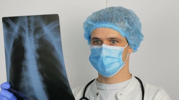 Młody chirurg w ochronnej masce medycznej bada prześwietlenie płuc pacjenta i rozważa diagnozę. — Wideo stockowe