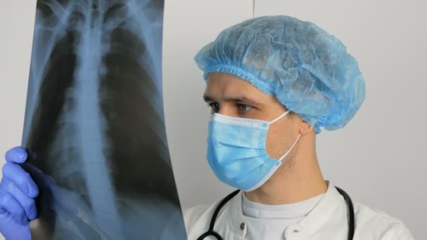 保護医療マスクを着用した若い外科医が肺のX線を検査し、診断を熟考します。彼の中に肺のX線写真を保持しています。 — ストック動画