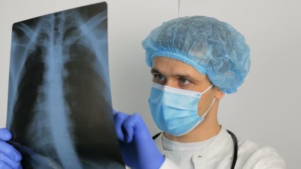 Egy fiatal sebész védőmaszkot visel, megvizsgálja a beteg tüdejének röntgenjét, és elgondolkodik a diagnózison. Egy fiatal, jóképű orvos orvosi maszkban röntgenképet tart a tüdejéről. — Stock videók