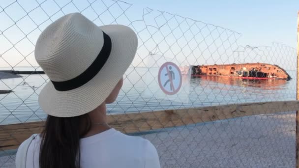 Beyaz şapkalı genç çekici kadın, deniz kıyısında yatan batık tankere üzülerek bakıyor. Çevre felaketi. Beyaz şapkalı güzel bir kadın batık petrol tankerine bakıyor.. — Stok video