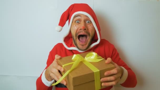 Ein junger gutaussehender Kerl in einem roten Weihnachtsmann-Kostüm hält ein Neujahrsgeschenk in die Höhe und gratuliert zu den Feiertagen. Ein junger attraktiver, lustiger Kerl im Silvesteranzug, als Santas Assistent einen New York rollt — Stockvideo