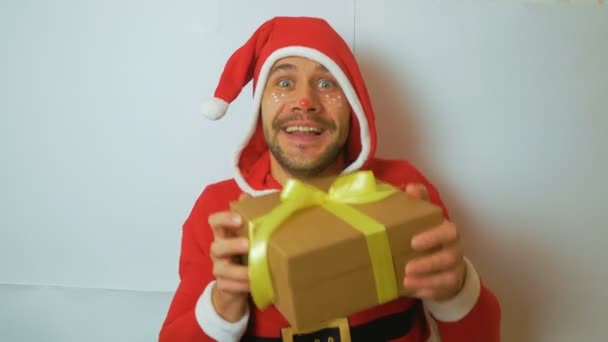 Un chico divertido atractivo joven en un traje de Año Nuevo como asistente de Santas rueda un regalo de Año Nuevo y se regocija en it.A chico guapo joven en un traje de carnaval santaclaus rojo sostiene un regalo de Año Nuevo — Vídeo de stock