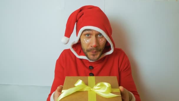 Młody przystojniak w czerwonym stroju karnawałowego Mikołaja wręcza prezent na Nowy Rok gratulując świąt. Młody zabawny facet w kostiumie elfa świątecznego uśmiecha się i macha ręką w powitaniu — Wideo stockowe