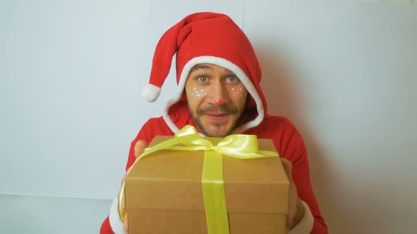 一个穿着红色桑塔克拉斯狂欢节服装的英俊小伙子献上了一份新年礼物，祝贺他节日快乐。一个年轻迷人的滑稽小伙子穿着新年套装当圣诞老人的助手翻开了一本新书。 — 图库视频影像
