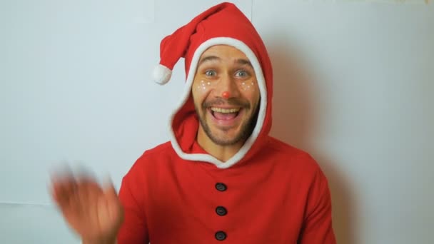 Młody, zabawny facet w kostiumie elfa z Nowego Roku uśmiecha się i macha ręką w powitaniu. Młody przystojniak w czerwonym stroju karnawałowego Mikołaja kręci głową i uśmiecha się szeroko. — Wideo stockowe