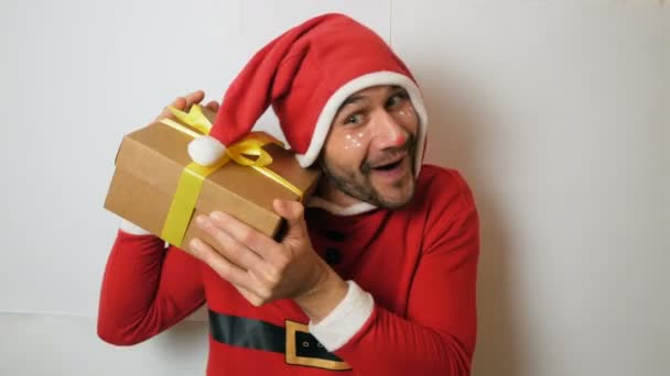 Um cara engraçado atraente jovem em um terno de Ano Novo como assistente de Santas rola um presente de Ano Novo e se alegra com ele.Um cara bonito jovem em um traje de carnaval santaclaus vermelho mantém um presente de Ano Novo — Vídeo de Stock