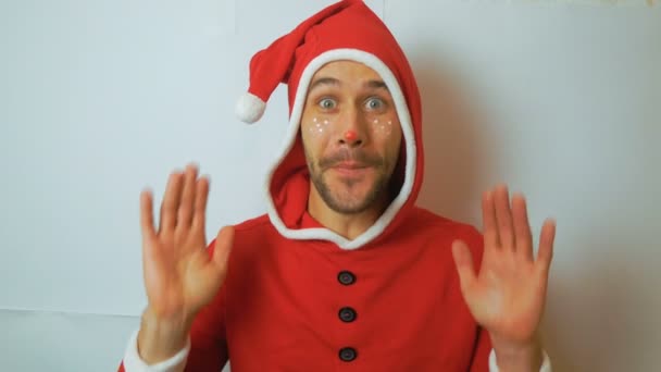 A young funny guy in a New Years elf costume smiles and waves his hand in greeting. A young handsome guy in a red santaclaus carnival costume shakes his head and smiles broadly. — Stock Video