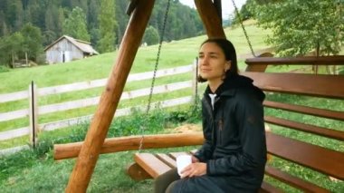 Genç ve çekici bir çiftçi kadın, ahşap salıncakta sallanırken kahve içiyor. Genç ve çekici bir kadın kahve içiyor ve dağların ve tarlanın güzel manzarasının tadını çıkarıyor. Genç ve sevimli.