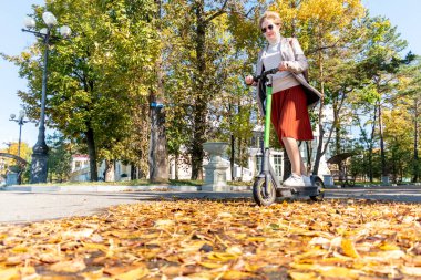 Şehirde elektrikli scooter kullanan orta yaşlı bir kadın. Modern teknolojiyi kullanma kavramı. Kısa kızıl saçlı insanlar. Sonbahar zamanı.