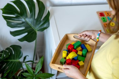 Çocuk raftan bir tepsi Montessori malzemesi alıyor. Evde eğitim kavramı. Tahta küplerden yapılmış oyuncaklar.