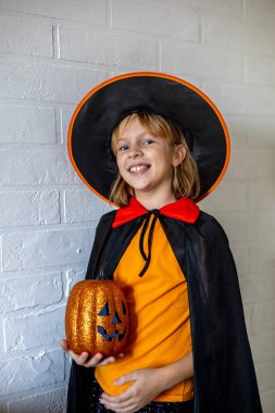 Cadı kostümü giymiş bir çocuk elinde dekoratif bir balkabağı tutuyor. Kötülük güler. Cadılar Bayramı konsepti. Boşluğu kopyala.