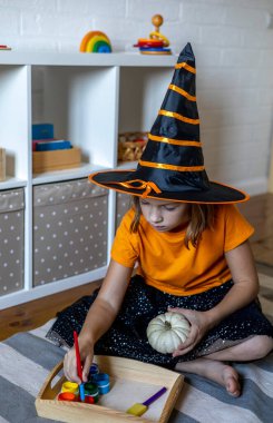 Cadı gibi giyinmiş bir kız fırçayı boyayla hamile bırakıyor. Cadılar bayramı için bir balkabağı yap. Çocuk oyunu konsepti. Evde eğitim. Montessori malzemesi