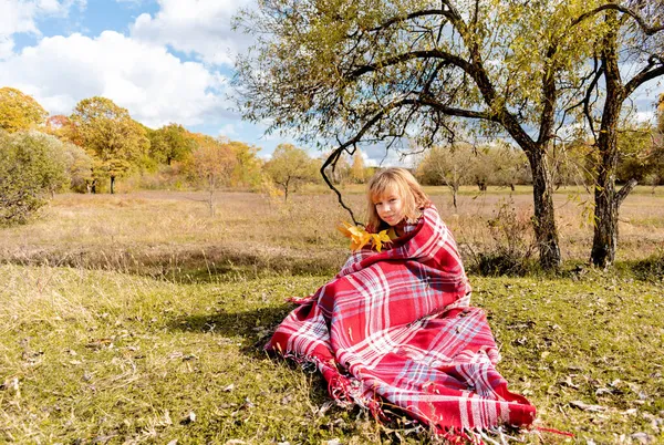 身穿黄色针织毛衣 手持树叶的少女坐在毛毯上 舒适缓慢的生活方式概念 在树旁的草地上野餐 秋天用热饮热身 — 图库照片