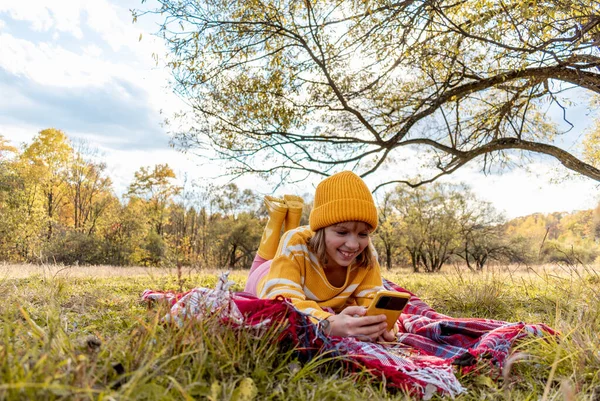 快乐的女孩用他的智能手机摄像头拍下了视频 舒适缓慢的生活方式概念 在树旁的草地上野餐 秋天用热饮热身 — 图库照片