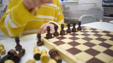 Çizgili sarı kazaklı kız tahta satranç tahtasında satranç oynuyor ve elinde satranç figürleri var. Boş zaman ve hobi anlayışı. Okul satranç kulübüne giden bir çocuk. Eğitim ve Geliştirme.