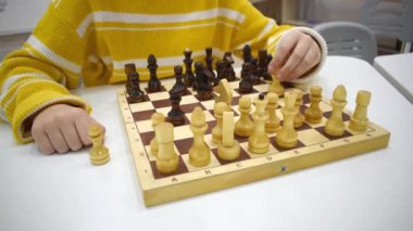 Çizgili sarı kazaklı kız tahta satranç tahtasında satranç oynuyor ve elinde satranç figürleri var. Boş zaman ve hobi anlayışı. Okul satranç kulübüne giden bir çocuk. Eğitim ve Geliştirme.