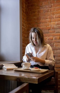 Genç bir kadın kahve içerken cep telefonundan mesaj yazıyor. İş kadını sosyal mesafeyi koruyor ve uzaktan çalışıyor. Yeni normal kavram