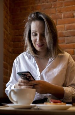 Genç bir kadın kahve içerken cep telefonundan mesaj yazıyor. İş kadını sosyal mesafeyi koruyor ve uzaktan çalışıyor. Yeni normal kavram