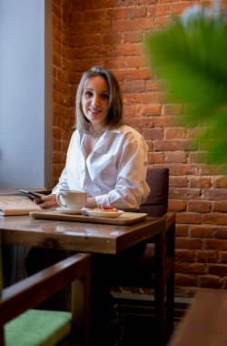 Genç bir kadın kahve içerken cep telefonundan mesaj yazıyor. İş kadını sosyal mesafeyi koruyor ve uzaktan çalışıyor. Yeni normal kavram