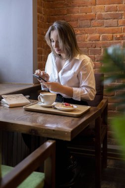 Genç bir kadın kahve içerken cep telefonundan mesaj yazıyor. İş kadını sosyal mesafeyi koruyor ve uzaktan çalışıyor. Yeni normal kavram