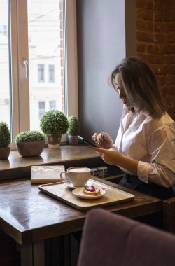 Genç bir kadın kahve içerken cep telefonundan mesaj yazıyor. İş kadını sosyal mesafeyi koruyor ve uzaktan çalışıyor. Yeni normal kavram