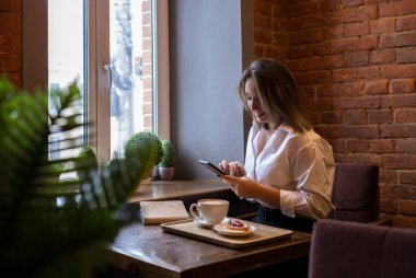 Genç bir kadın kahve içerken cep telefonundan mesaj yazıyor. İş kadını sosyal mesafeyi koruyor ve uzaktan çalışıyor. Yeni normal kavram