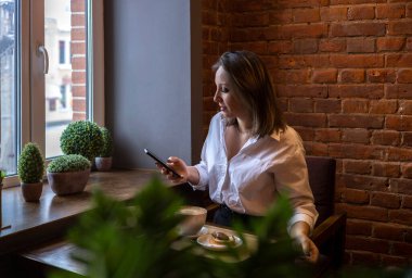 Genç bir kadın kahve içerken cep telefonundan mesaj yazıyor. İş kadını sosyal mesafeyi koruyor ve uzaktan çalışıyor. Yeni normal kavram