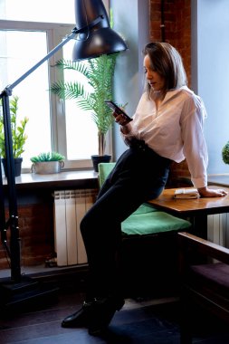 Genç bir kadın akıllı telefonu elinde tutuyor, pencerenin yanında duruyor. Teneffüste mahremiyet. İş görüşmesi