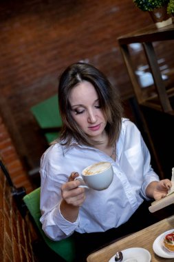 Beyaz gömlekli genç bir kadın kafede oturuyor ve elinde bir fincan kahve tutuyor. Yemek molası. Ofis çalışanı kahvaltı yapıyor.