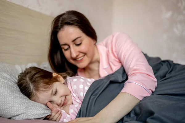 パジャマを着た美しい母親はベッドで寝ていて 小さな娘を抱きかかえています 幸せな家族だ ゆっくりと朝だ 母の日 — ストック写真