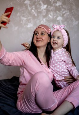 Pijamalı mutlu anne ve kız yatakta akıllı telefonlu selfie çekiyor. Aile kavramı ve Anneler Günü kavramı